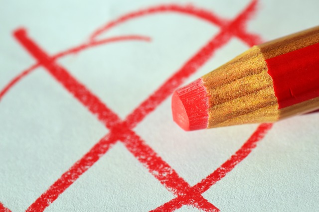 Nahaufnahme: Stumpfe Spitze eines roten Buntstiftes vor einem Blatt Papier, darauf wurde ein Kreis mit einem X durch gemalt.