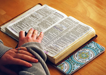 Aufgeschlagene Bibel auf hellbraunem Holztisch. Darunter Notizbuch mit Muster aus kreisförmig angeordneten Flecken. Hände.