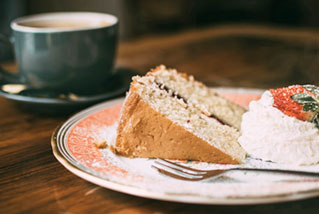 Türkise Kaffeetasse auf dunklem Holz, weiß-rötlicher Teller mit Goldrand, darauf Kuchenstück, Schlagobers, Erdbeere, Gabel.