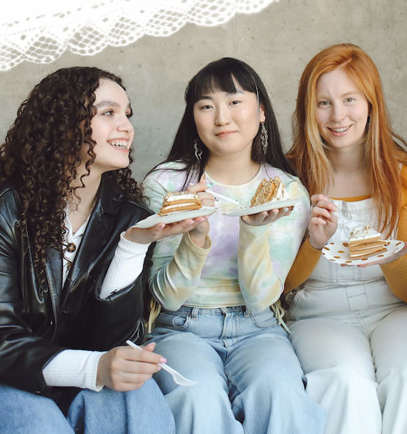 Drei Mädchen essen mehrlagigen Kuchen. Eins hat braune Locken, eines wirkt asiatisch mit schwarzem Haar, eines hat rote Haare.