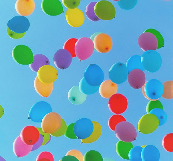 Viele Luftballons vor blauem Himmel: mittel- u. hellbau, hell- u. dunkelgrün, lila, rosa, rot, gelb, orange. Viele überlappen.