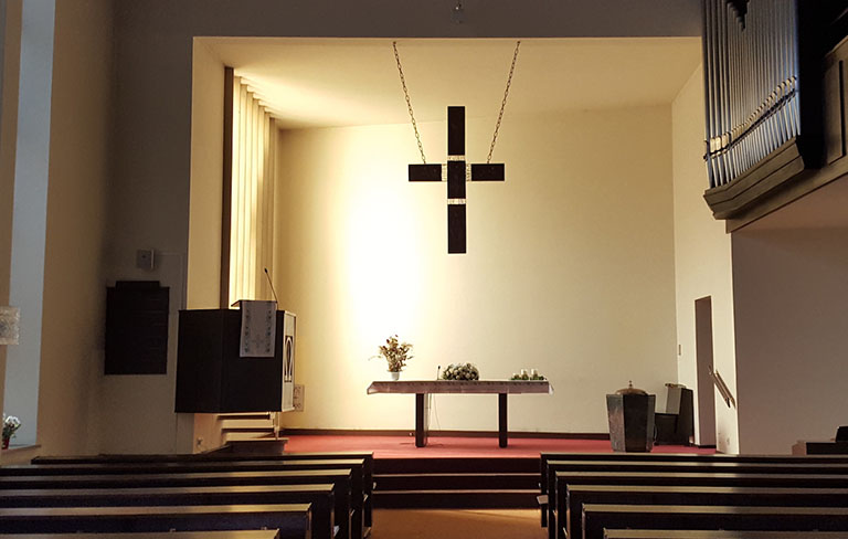 Der Kirchenraum, helles weißes Licht in der Mitte, dunkelbraunes Kreuz, Empore mit rotem Boden, Bänke, Blumen, Orgelpfeifen.