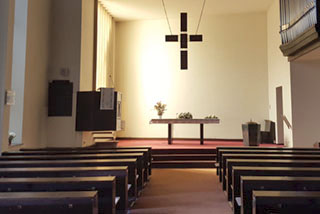 Der Kirchenraum, helles weißes Licht in der Mitte, dunkelbraunes Kreuz, Empore mit rotem Boden, Bänke, Blumen, Orgelpfeifen.